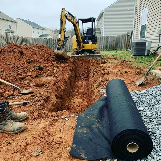 Freshly excavated trench for French drain installation with mini excavator, rolled geotextile fabric and pile of gravel staged nearby